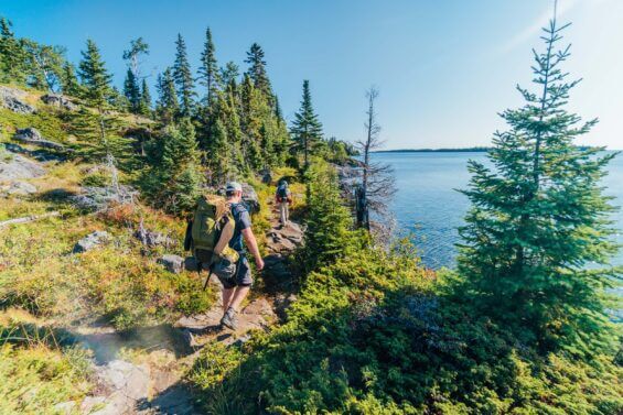 Two hikers with backpacks full of gear walk on a cliff-side trail in Isle Royale National Park.
