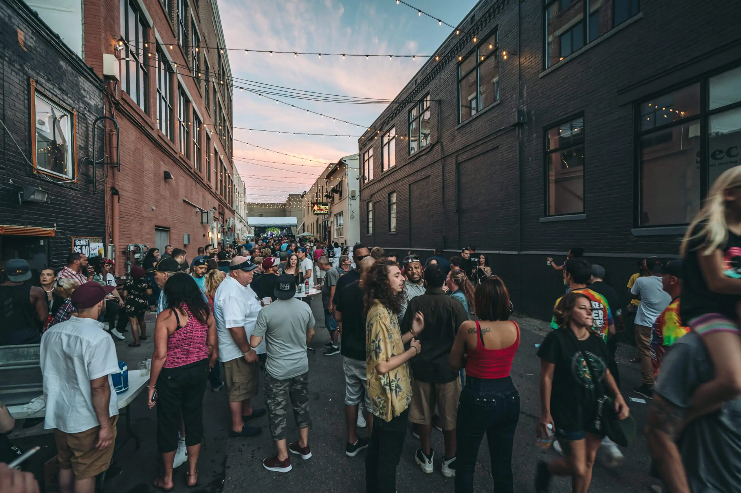 Groups gather downtown in an alley lit by string lights between two brick buildings for Alley Fest in Flint.
