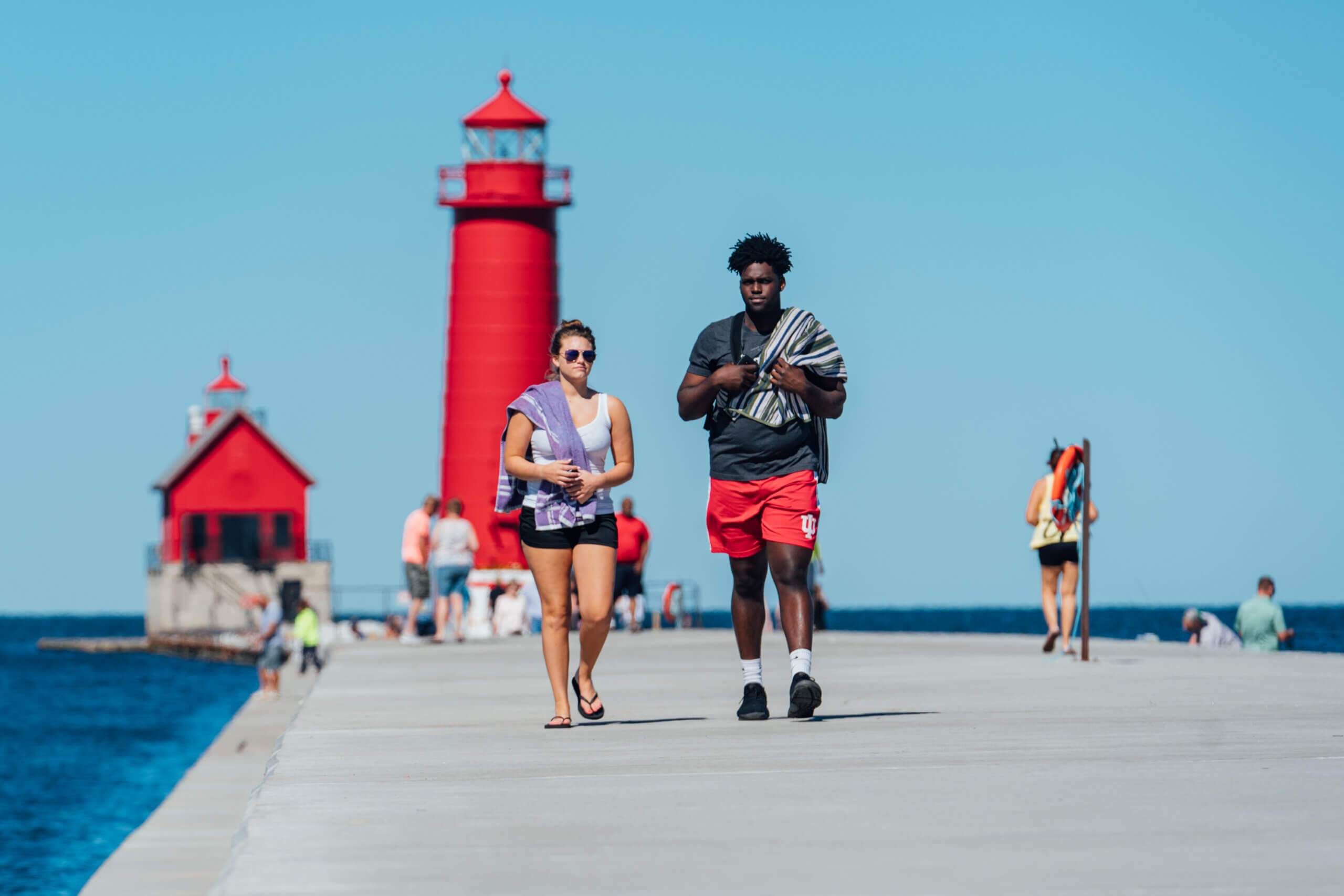 Two people with beach towels walk away from the bright red Grand Haven lighthouse.