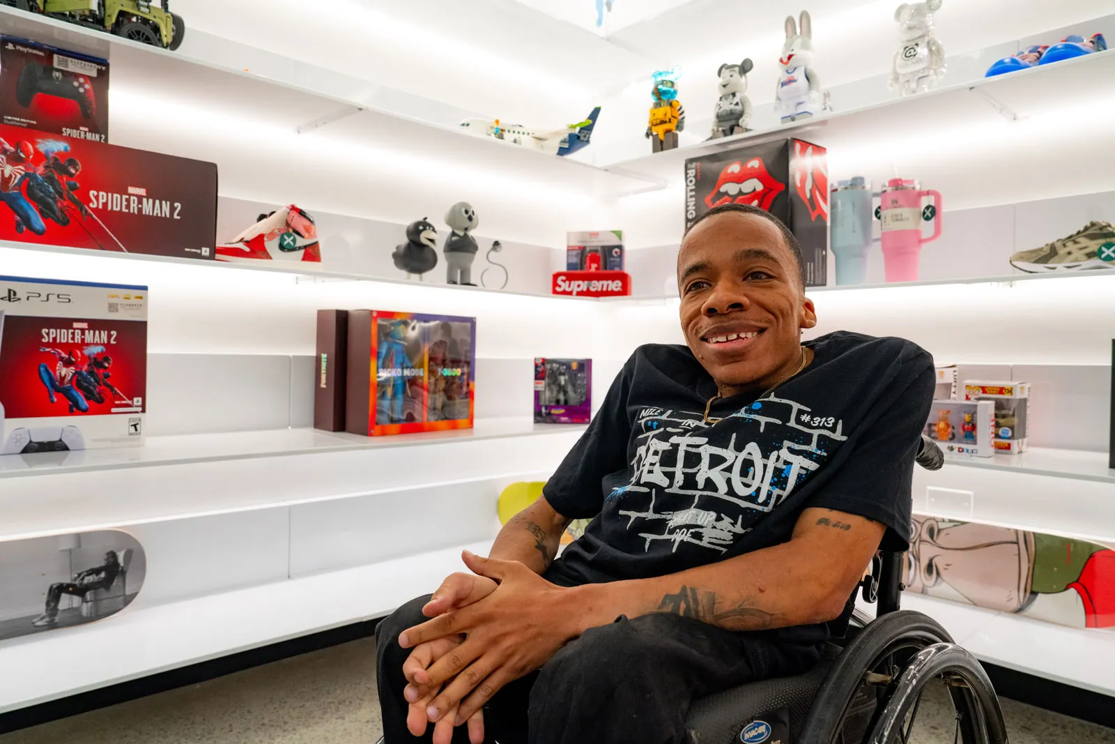 Devin Allen smiles warmly from his wheelchair while sitting in front of white shelves adorned with sports and media memorabilia.