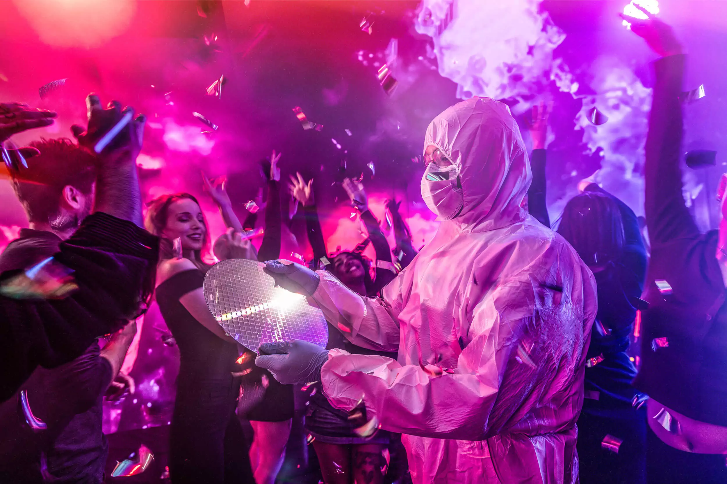Engineer in white, full-body, protective lab gear, clear goggles, a surgical mask and latex gloves holds a circular semiconductor plate in a crowd of partying people with shining confetti falling.