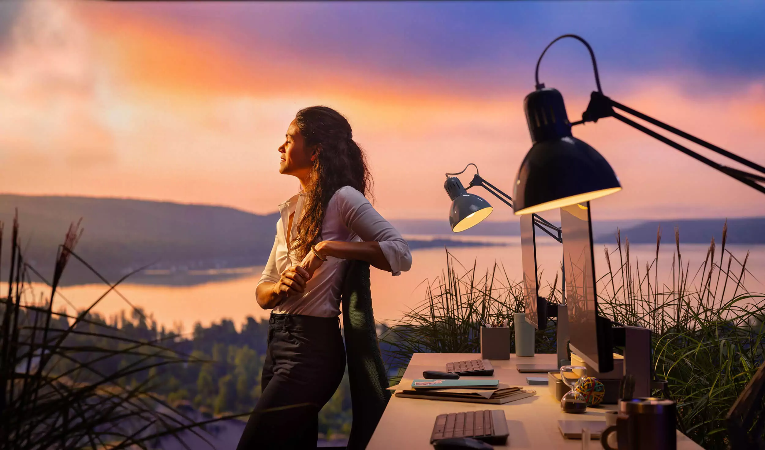 A woman in professional dress leans against an office chair in front of an organized desk with sunlight beaming on her face juxtaposed over a stunning lake scene at sunset.