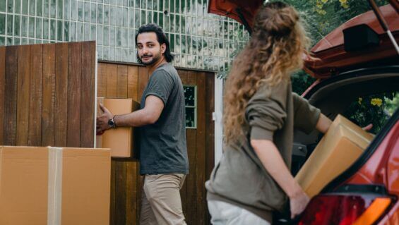 Husband smiles back at wife while carrying a moving box into their new house.