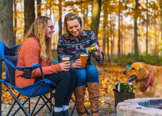 Two women smile, enjoying IPAs outdoors in fold-up chairs amidst the beautiful fall foliage.